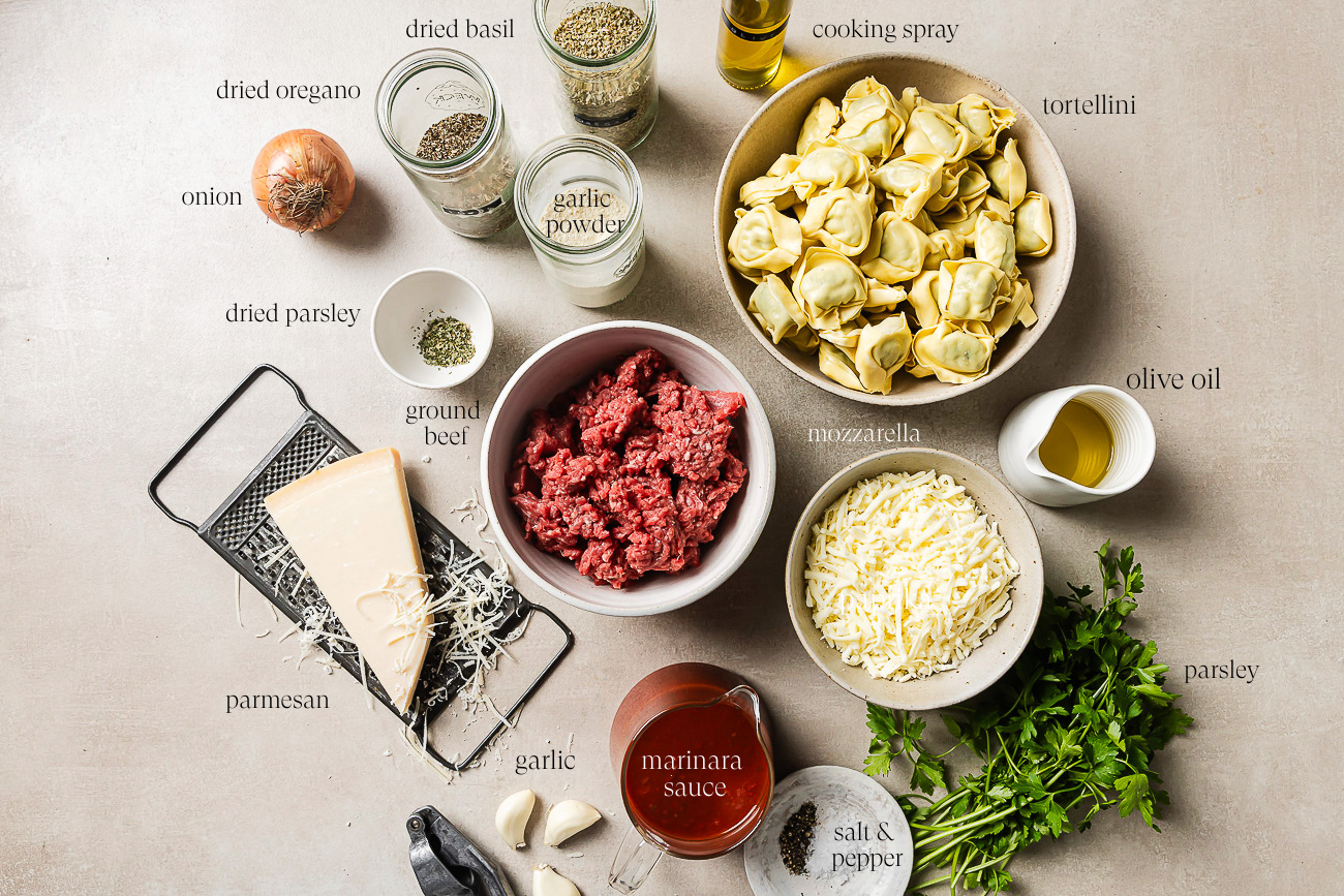 Ingredients for Baked Tortellini with Ground Beef are arranged on a counter, including tortellini, ground beef, mozzarella, Parmesan, marinara sauce, fresh and dried herbs, onion, garlic, spices, olive oil, and cooking spray.
