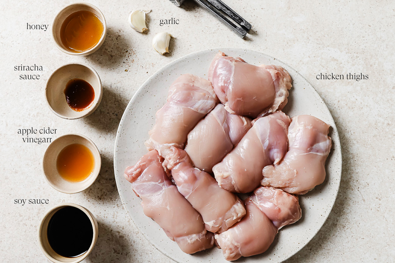 Raw chicken thighs on a plate, ready to make Honey Garlic Chicken Thighs, are surrounded by bowls of honey, sriracha sauce, apple cider vinegar, soy sauce, fresh garlic cloves, and measuring spoons on a light countertop.