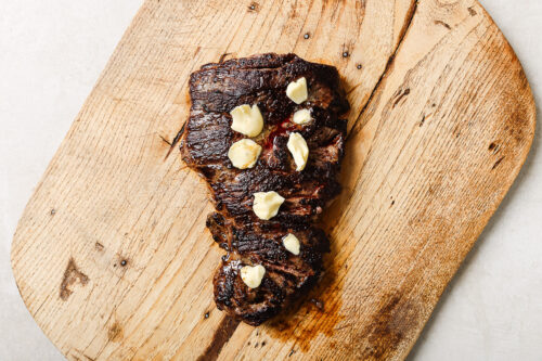 A pan seared flank steak topped with dollops of melting butter sits on a rustic wooden cutting board.