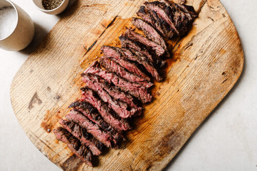 Pan Seared Flank Steak sliced medium-rare and arranged neatly on a rustic wooden cutting board, with a bowl of pepper and a bowl of coarse salt nearby.