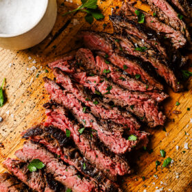 Sliced medium-rare steak garnished with parsley on a wooden board, with a bowl of coarse salt and scattered salt crystals nearby.