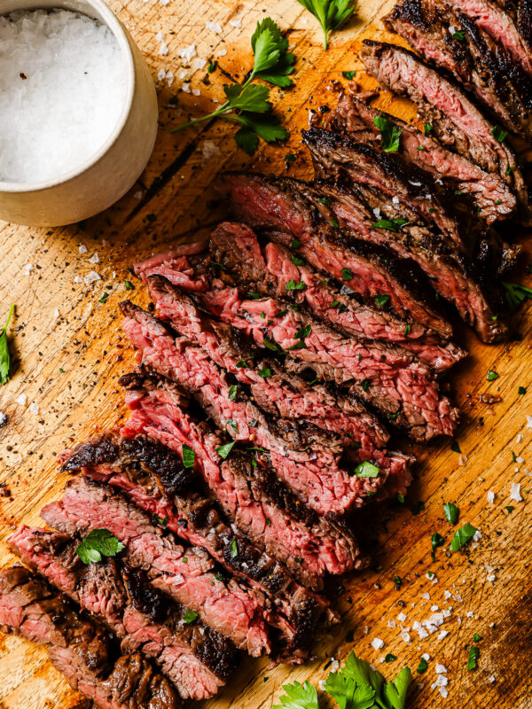 Sliced medium-rare steak garnished with parsley on a wooden board, with a bowl of coarse salt and scattered salt crystals nearby.
