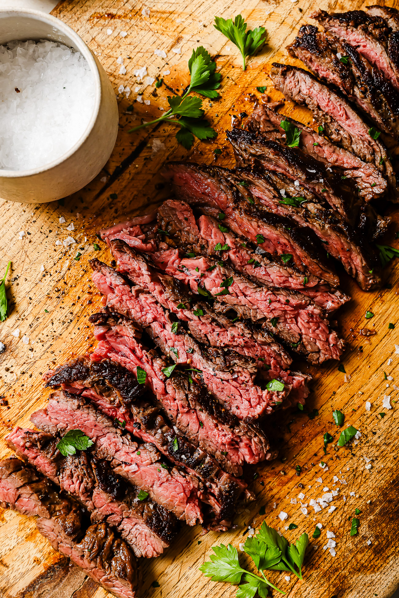 Sliced medium-rare steak garnished with fresh parsley on a wooden cutting board, with coarse salt scattered around and a small bowl of salt nearby.