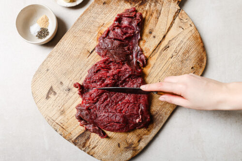 A hand slices raw flank steak on a wooden cutting board, preparing it for a delicious pan seared flank steak. Nearby, a small bowl contains salt and pepper.