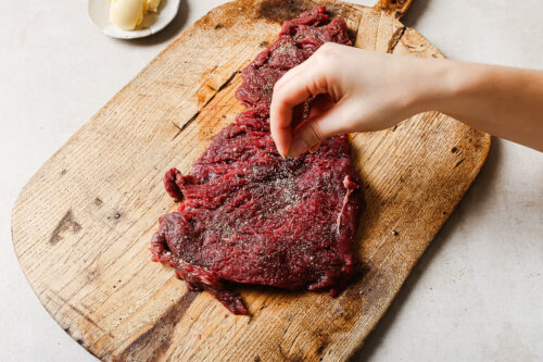 A hand sprinkles seasoning onto a raw skirt steak placed on a wooden cutting board, ready to become a delicious Pan Seared Flank Steak. A small dish with a pat of butter sits in the background.