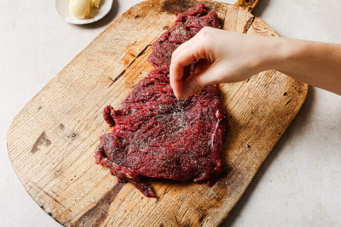 A hand sprinkles seasoning onto a raw skirt steak placed on a wooden cutting board, ready to become a delicious Pan Seared Flank Steak. A small dish with a pat of butter sits in the background.