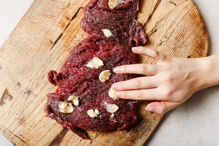 A hand rubs slices of garlic and seasoning onto a raw Pan Seared Flank Steak placed on a wooden cutting board.