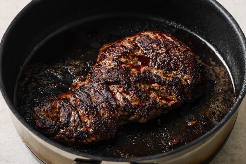 A large Pan Seared Flank Steak is cooking in a black skillet, displaying a dark brown, caramelized crust with flavorful juices pooling around it.