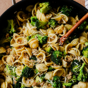 A skillet of Orecchiette with Sausage and Broccoli is topped with grated cheese, a wooden spoon nestled inside. A white cloth rests nearby. Text reads Orecchiette with Sausage and Broccoli.