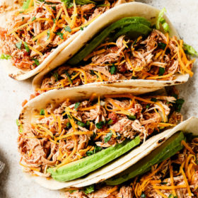 Overhead view of four Slow Cooker Chicken Tacos filled with shredded chicken, cheese, avocado slices, and chopped herbs on a light surface. Text above reads “Slow Cooker Chicken Tacos.”.