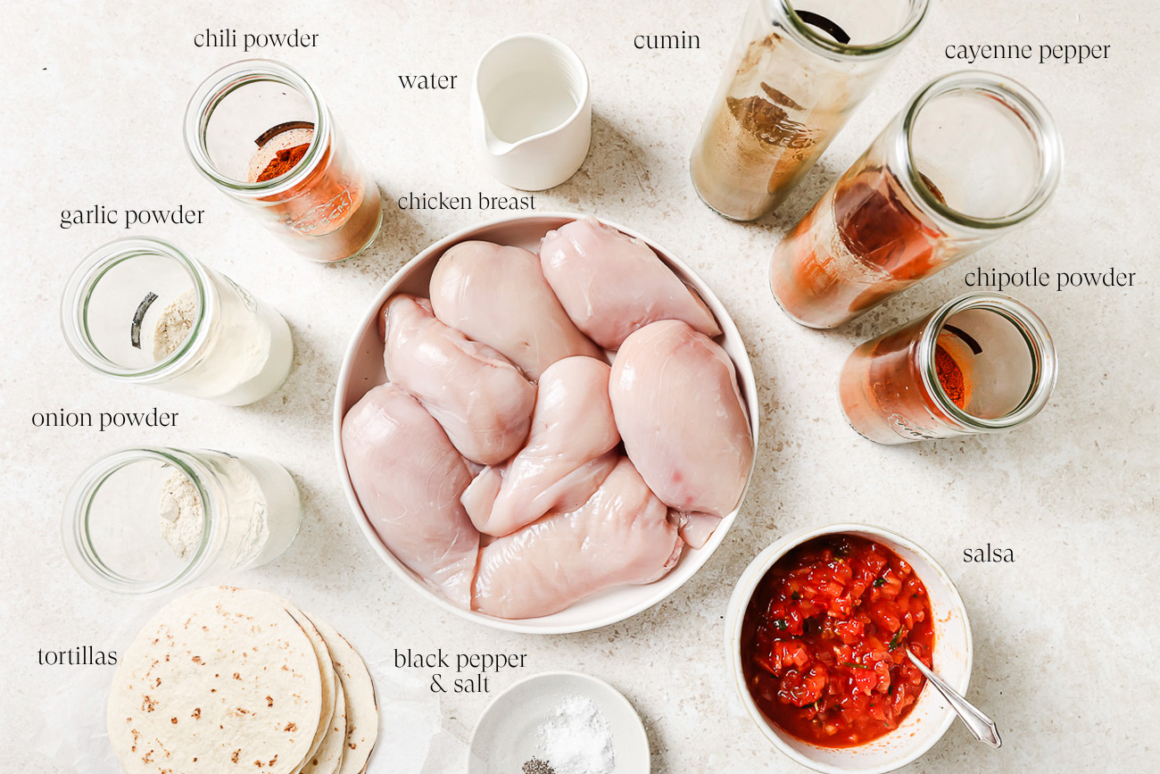 A bowl of raw chicken breasts surrounded by labeled jars of spices, a cup of water, salsa, tortillas, salt, and black pepper—perfect ingredients for making flavorful Slow Cooker Chicken Tacos.
