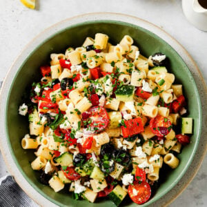 A green bowl filled with pasta salad containing cherry tomatoes, cucumbers, red peppers, black olives, feta cheese, and chopped herbs. Small bowls of salt and chopped herbs, lemon wedges, and a napkin are nearby.