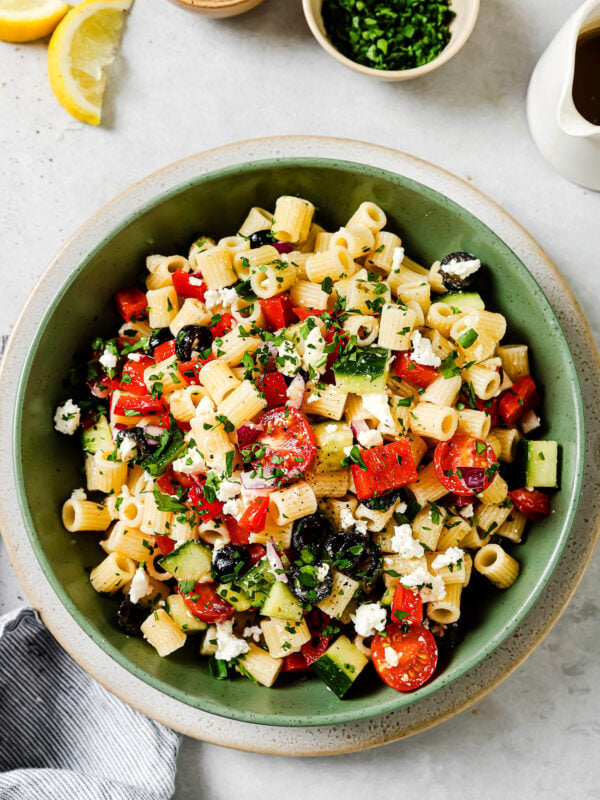 A green bowl filled with pasta salad containing cherry tomatoes, cucumbers, red peppers, black olives, feta cheese, and chopped herbs. Small bowls of salt and chopped herbs, lemon wedges, and a napkin are nearby.