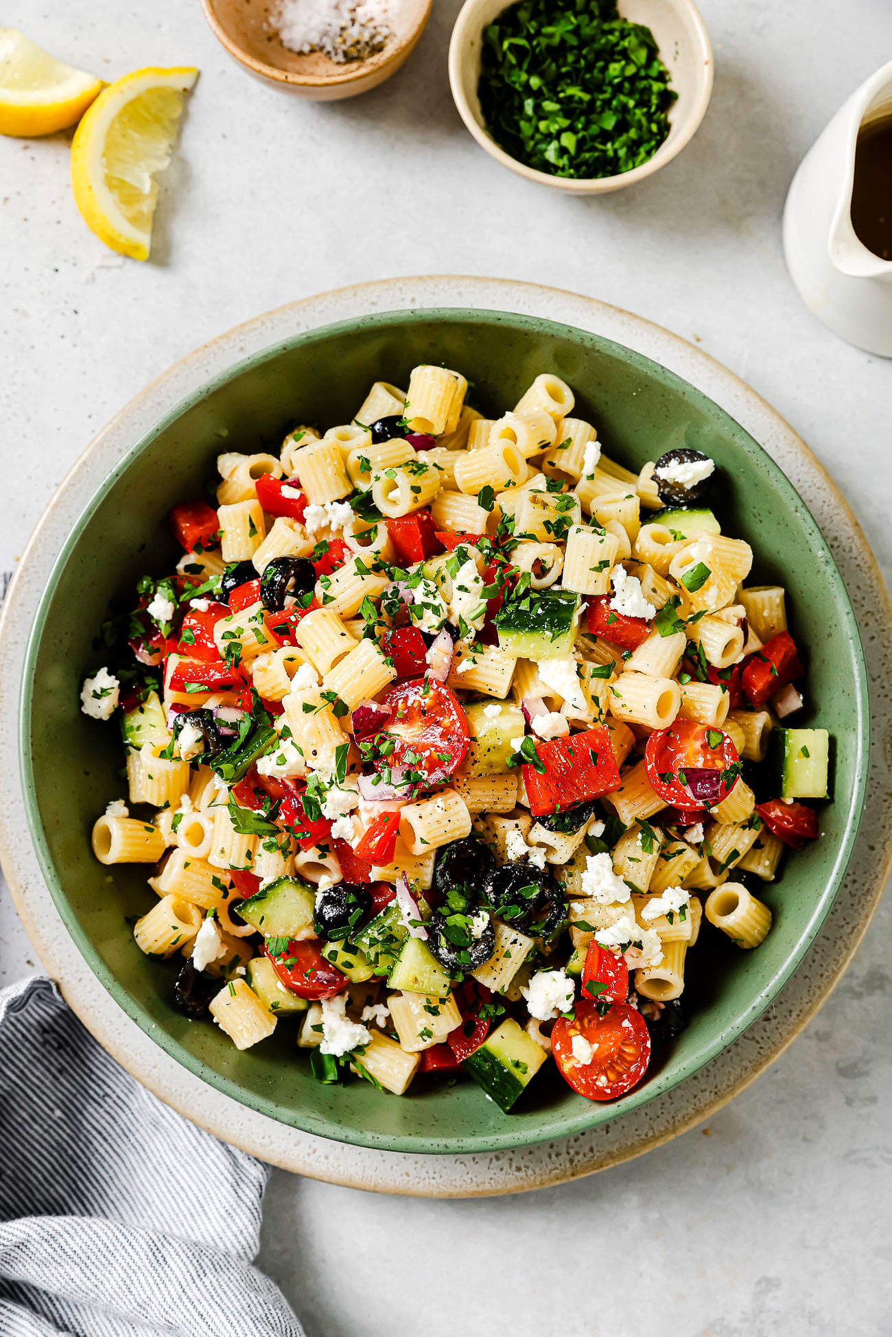 A green bowl filled with pasta salad containing cherry tomatoes, cucumbers, red peppers, black olives, feta cheese, and chopped herbs. Small bowls of salt and chopped herbs, lemon wedges, and a napkin are nearby.