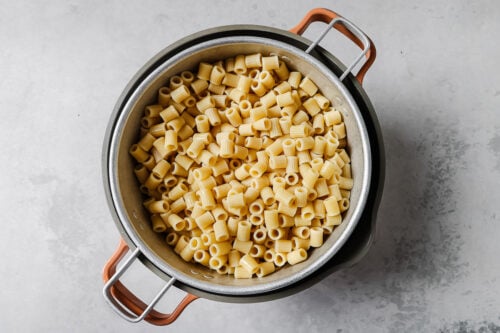 A colander filled with cooked rigatoni pasta sits atop a pot with brown handles on a light gray surface.