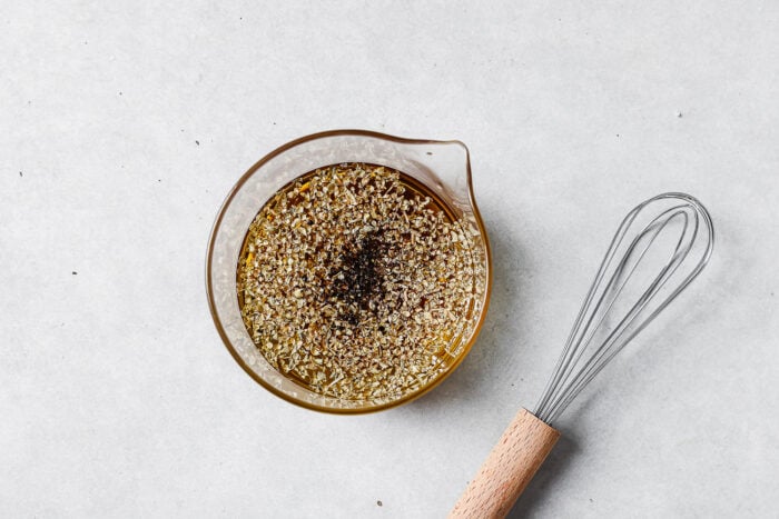 A glass measuring cup filled with oil and herbs sits on a light surface next to a metal whisk with a wooden handle.
