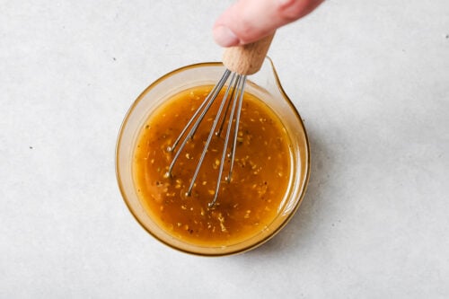 A hand holding a whisk is mixing a yellow-brown liquid with herbs in a small glass bowl on a light gray surface.