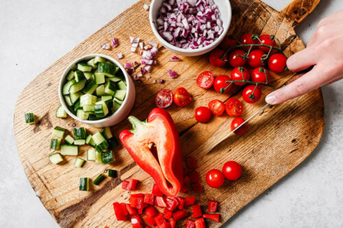 A hand slices cherry tomatoes on a wooden cutting board, surrounded by chopped cucumber, red bell pepper, diced red onion, and a bowl of whole and halved cherry tomatoes.