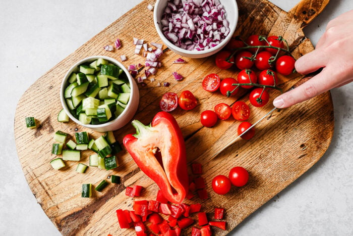 A hand slices cherry tomatoes on a wooden cutting board, surrounded by chopped cucumber, red bell pepper, diced red onion, and a bowl of whole and halved cherry tomatoes.