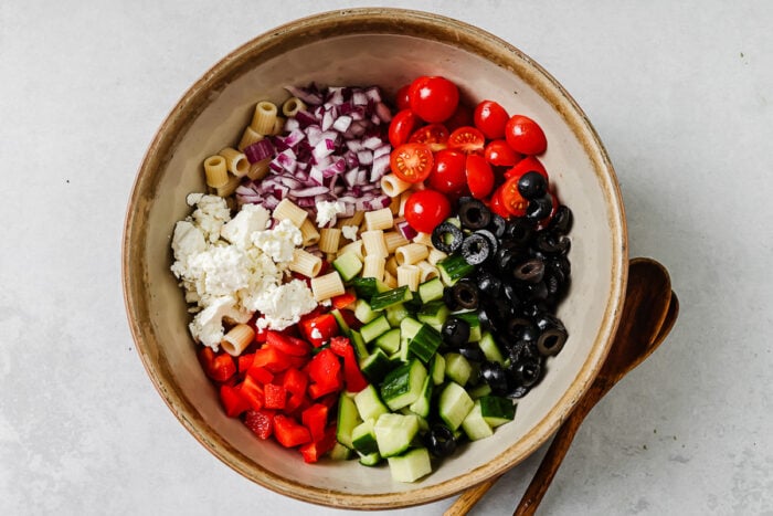 A large bowl containing cooked pasta, chopped red onions, cherry tomatoes, sliced black olives, cucumber, diced red bell pepper, and crumbled feta cheese, ready to be mixed into a salad. Wooden salad servers lie beside the bowl.