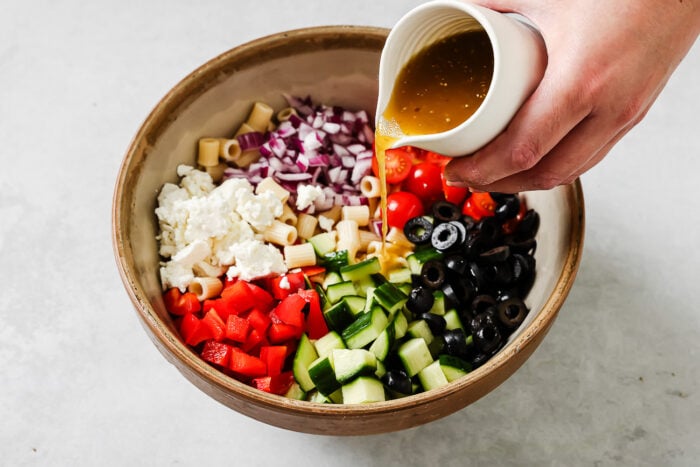 A hand pours dressing from a small white pitcher onto a bowl filled with chopped cucumbers, red bell peppers, cherry tomatoes, black olives, red onions, pasta, and crumbled feta cheese.