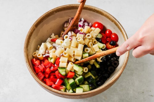 A person uses wooden utensils to mix a bowl filled with pasta, chopped red bell pepper, cucumber, cherry tomatoes, red onion, black olives, and feta cheese.