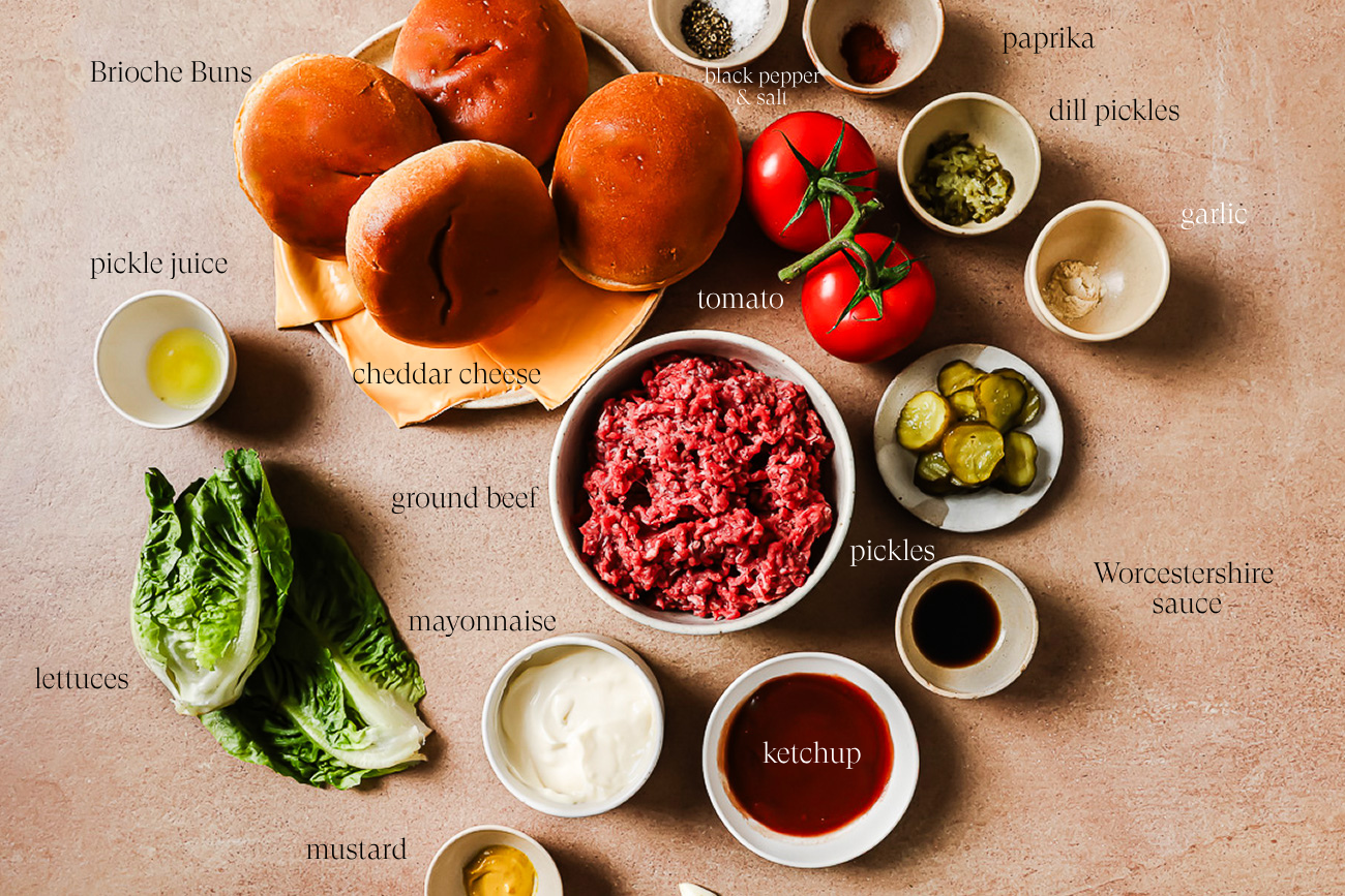 A flat lay of Smash Burger Recipe ingredients including brioche buns, cheddar cheese, tomato, ground beef, pickles, lettuce, mayonnaise, mustard, ketchup, Worcestershire sauce, garlic, dill pickles, pickle juice, and seasonings.