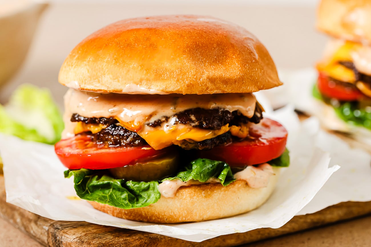 A close-up of a cheeseburger with two beef patties, melted cheddar cheese, lettuce, tomato slices, pickles, and sauce in a shiny, toasted bun, placed on parchment paper on a wooden surface.