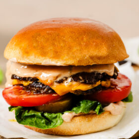A close-up of a juicy cheeseburger with a golden bun, lettuce, tomato slices, melted cheese, two beef patties, and creamy sauce, served on parchment paper atop a wooden board.