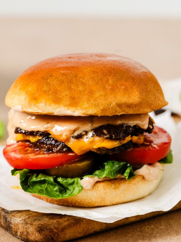 A close-up of a juicy cheeseburger with a golden bun, lettuce, tomato slices, melted cheese, two beef patties, and creamy sauce, served on parchment paper atop a wooden board.