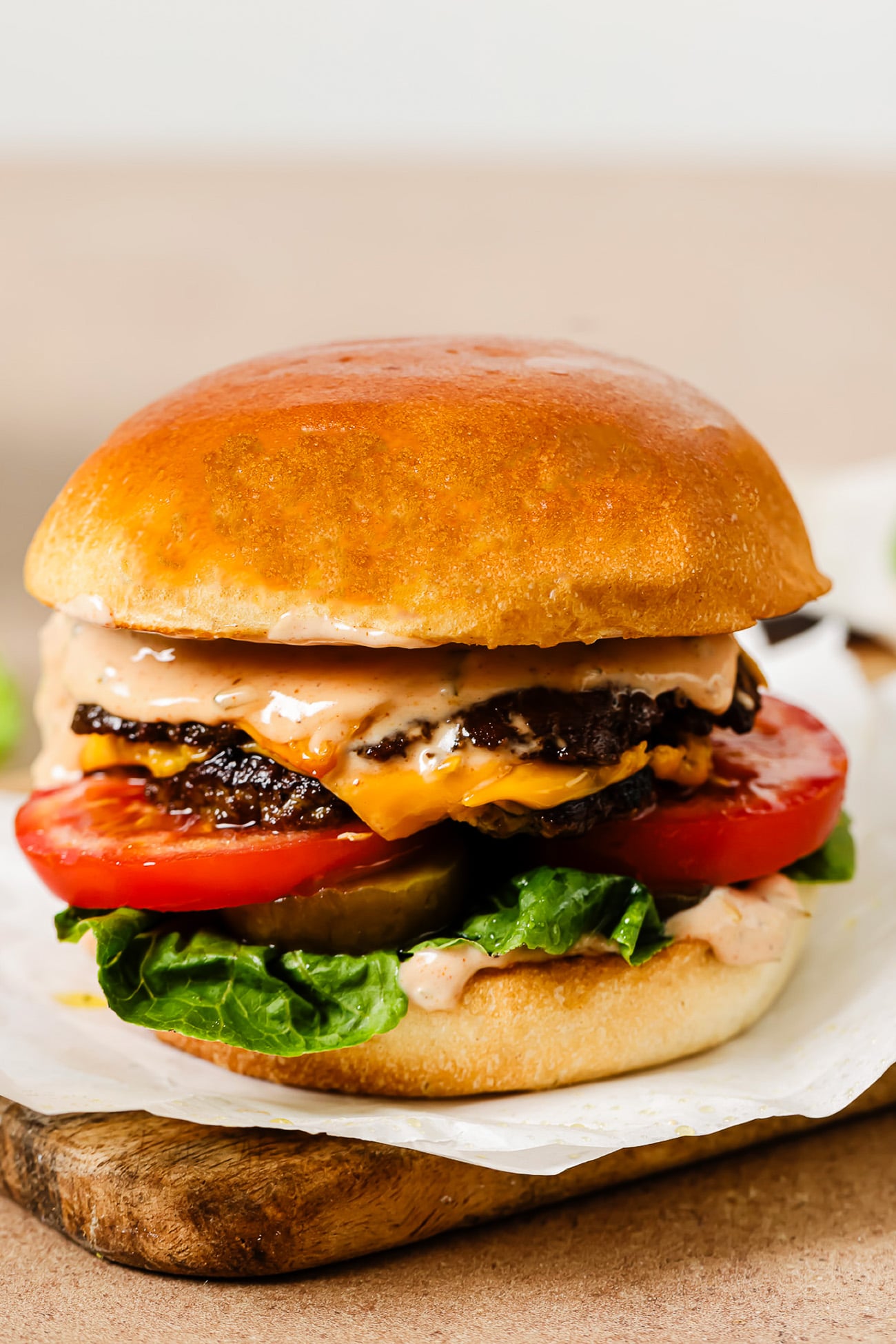 A close-up of a juicy cheeseburger with a golden bun, lettuce, tomato slices, melted cheese, two beef patties, and creamy sauce, served on parchment paper atop a wooden board.