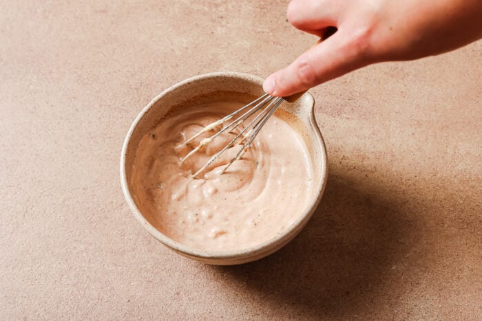 A hand whisking a creamy, light brown sauce or dressing in a beige ceramic bowl on a light brown surface.