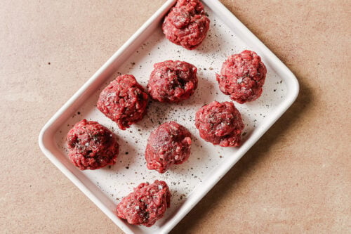 A white rectangular tray holds eight raw beef meatballs sprinkled with ground black pepper, set on a light brown surface.