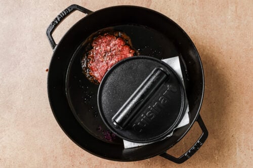 A raw burger patty is being pressed with a weighted lid on top, cooking in a black cast iron skillet on a light brown surface.