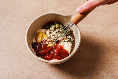A hand uses a whisk to mix ingredients including ketchup, mayonnaise, mustard, relish, and seasonings in a small beige bowl on a tan countertop.