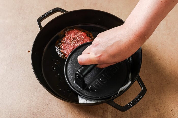 A hand presses a black cast iron burger press onto a raw beef patty in a black cast iron skillet on a tan surface.