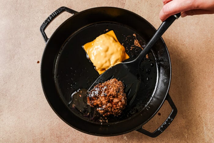 A hand uses a spatula to lift a cooked hamburger patty in a black skillet. Another patty with a slice of melted yellow cheese is also in the skillet on a beige countertop.