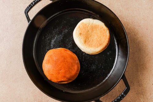 A cast iron skillet with two English muffins inside, one with a golden brown top and the other with a lighter, pale top, sitting on a beige surface.