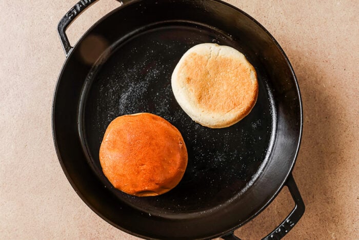 A cast iron skillet with two English muffins inside, one with a golden brown top and the other with a lighter, pale top, sitting on a beige surface.