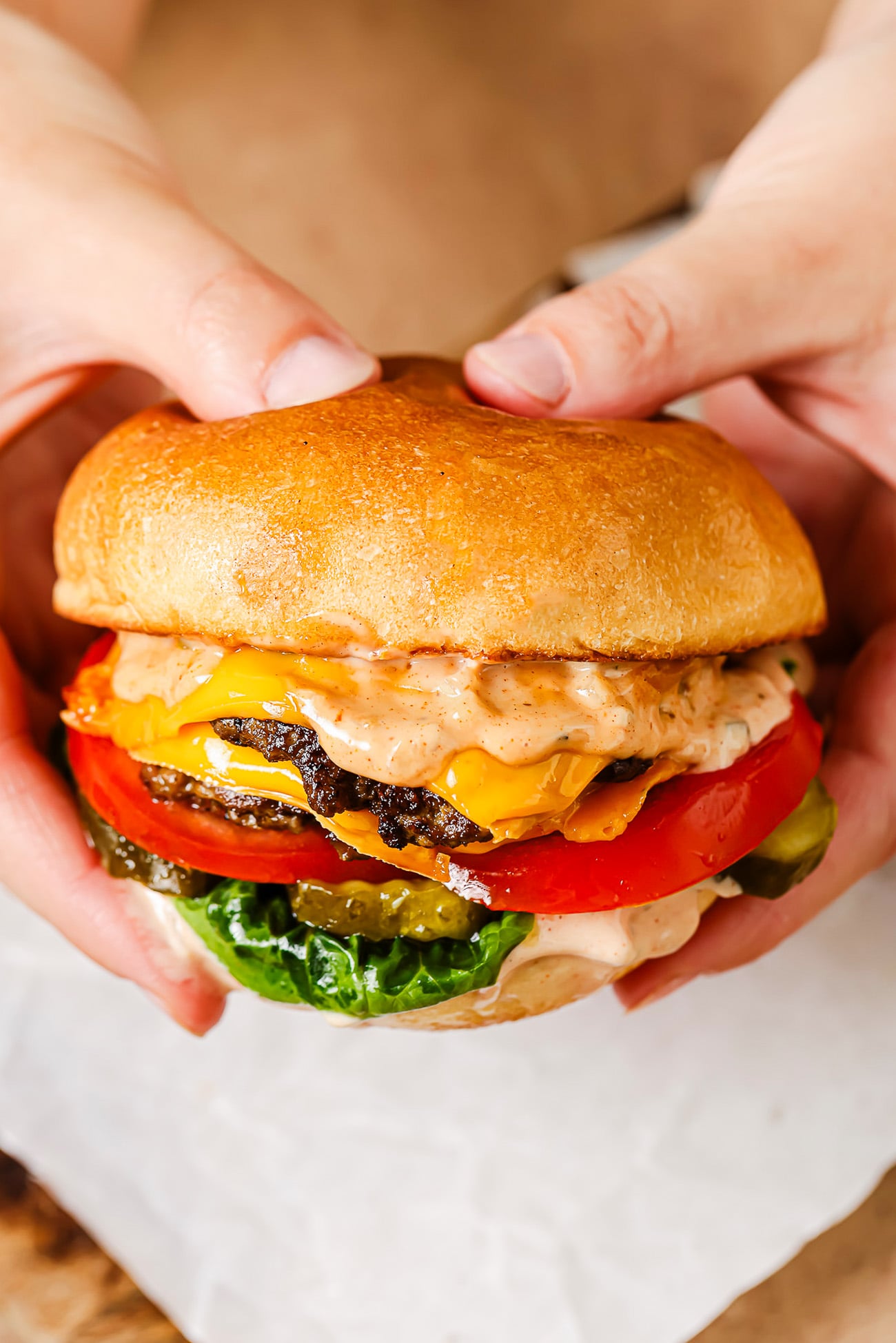 Two hands hold a burger with lettuce, pickles, tomato, cheddar cheese, a beef patty, and a generous layer of sauce between a golden-brown bun&mdash;an irresistible creation inspired by the classic Smash Burger recipe.