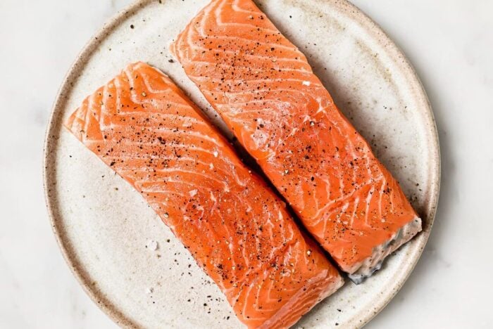 Two raw salmon fillets sprinkled with pepper sit side by side on a round, beige ceramic plate against a white background.