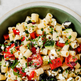 A green bowl filled with delicious Greek Pasta Salad featuring pasta, cherry tomatoes, cucumbers, olives, red peppers, feta cheese, and fresh herbs. Text above reads Easy and Quick Greek Pasta Salad.