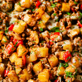 A skillet filled with a hearty Ground Beef and Potatoes mixture, diced red bell peppers, melted cheese, and fresh herbs, with a serving spoon scooping up some of the savory blend.