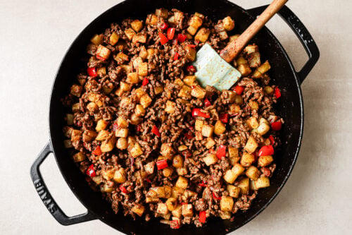 A skillet filled with Ground Beef and Potatoes, diced red bell peppers, all mixed together with a spatula for a hearty, flavorful meal.