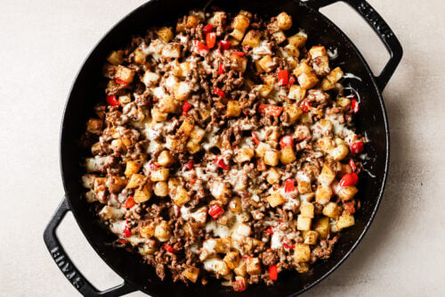 A black skillet filled with a savory Ground Beef and Potatoes mixture, combined with diced red bell peppers and melted cheese, sits invitingly on a light-colored surface.