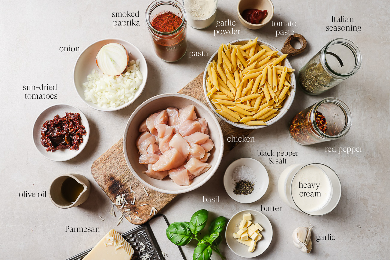 Ingredients for a Marry Me Chicken Pasta are arranged on a counter, including diced chicken, penne, cream, parmesan, herbs, spices, sun-dried tomatoes, onion, olive oil, butter, and garlic. Labels identify each ingredient.