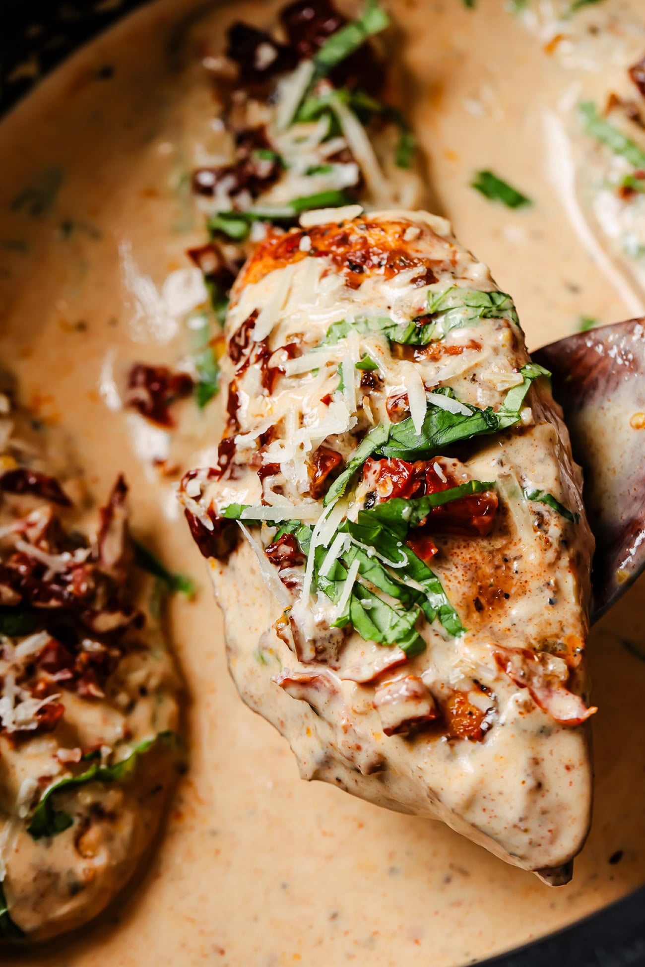 A chicken breast coated in a creamy sauce with sun-dried tomatoes, fresh basil, and grated cheese, lifted from a pan by a wooden spoon.