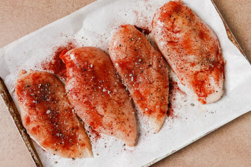 Four raw chicken breasts seasoned with red spice and black pepper on a parchment-lined baking sheet.