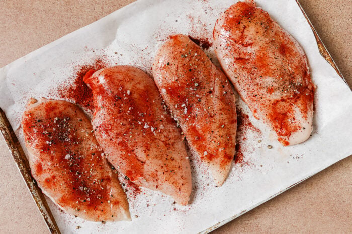 Four raw chicken breasts seasoned with red spice and black pepper on a parchment-lined baking sheet.