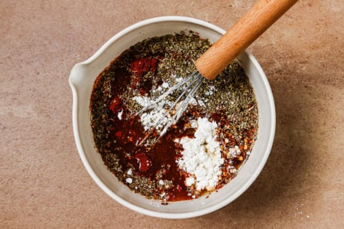 A wire whisk mixes oil, dried herbs, red spice, and a spoonful of white powder in a white bowl.